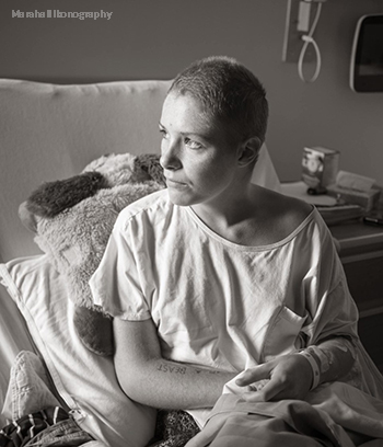 A young woman with a shaved head looks out a window, while sitting in her hospital bed.